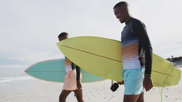 Happy african american teenage twin brothers walking on a beach carrying surfboards alt