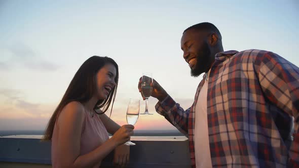 Multiracial Couple Laughing and Flirting, Clinking Champagne Glasses on Terrace