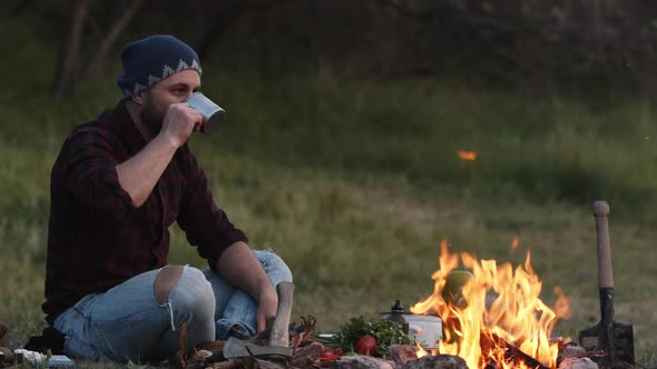 Man Sits Near Campfire and Drinks Hot Tea From Metal Tourist Cup Looks at Blazing Bonfire with