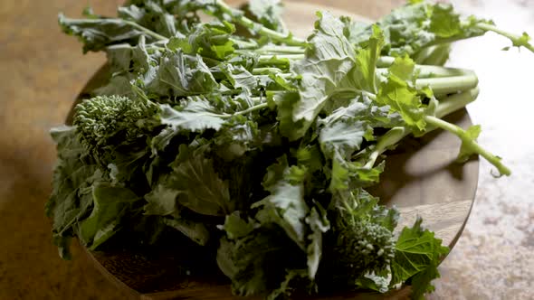 Clusters of stalks of fresh rapini on a round cutting board rotating in beautiful backlit light. alt