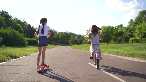 Back View Two Girls Riding Scooter And Bike On Driveway alt
