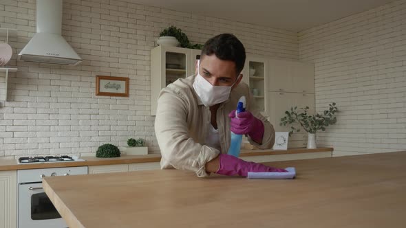 A handsome man in a protective mask and gloves wipes the table with a rag and disinfectant. alt