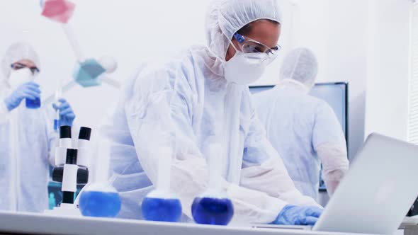 Focused Young Female Scientist in Coverall Protection Equipment Typing Report on Laptop alt