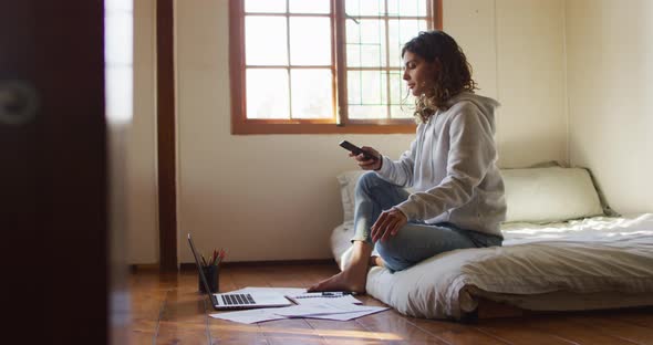 Mixed race woman working at home, sitting on bed using smartphone and laptop in cottage alt