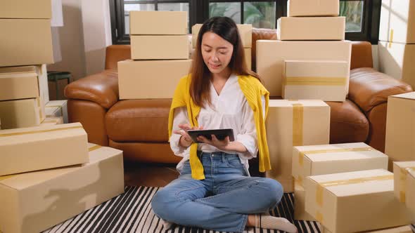 Wide shot of young Asian woman use tablet and sit among stack of carton or boxes