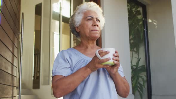 African american senior woman drinking coffee while standing at the front door at home alt