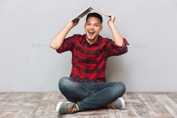 Cheerful young man in plaid sirt holding laptop over head