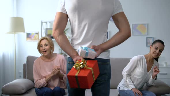Curious Wife and Mother-In-Law Looking at Man Holding Gift Boxes Behind Back alt