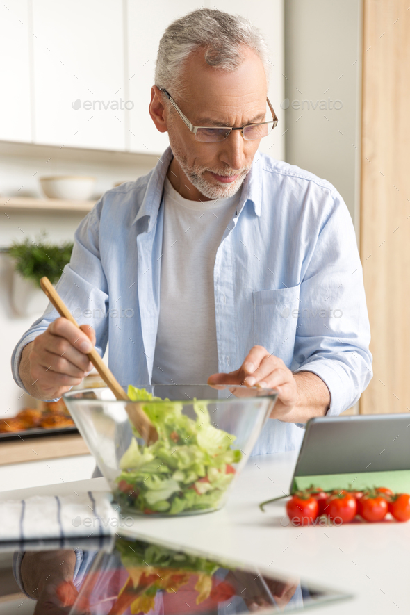 Handsome concentrated mature man cooking salad using tablet