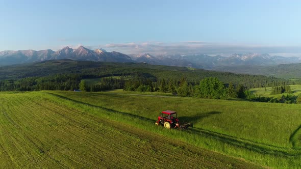 Tractor mowing the grass with beautiful high mountains in the background, aerial view.