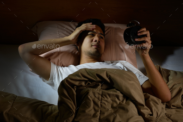 Depressed man suffering from insomnia looking at alarm clock lying in bed