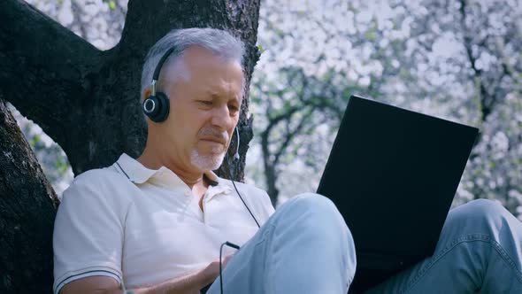 An Elderly Grayhaired Man with a Beard in Headphones and with a Laptop Works Online Sitting on the