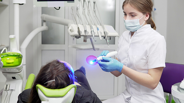 Young Female Dentist in Mask and Gloves Using Dental UV Light Equipment for Polymer Hardening alt