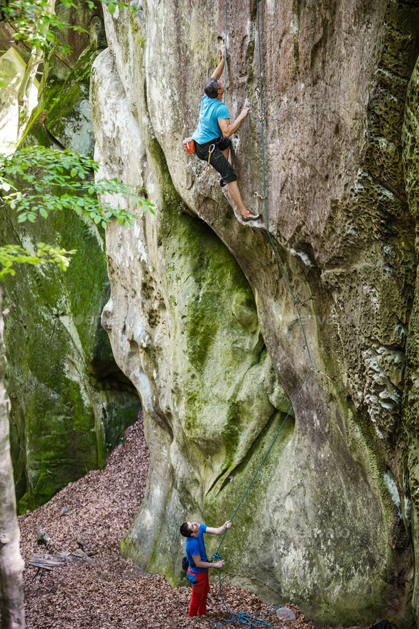 Young man climbing on vertical cliff, his partner belaying