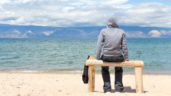 Man or Woman Is Sitting in a Hoodie on the Beach. Back View alt