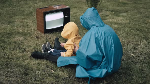 Woman with Small Child in Raincoats are Sitting on Grass and Watching an Old TV