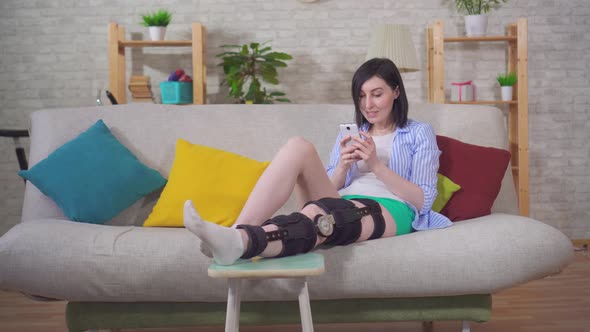 Female with the Phone After Injury Knee Bend Orthosis Knee Brace Lies on the Couch at Home alt