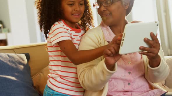 Smiling grandmother and granddaughter using digital tablet on sofa in living room 4k alt