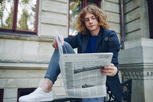 Young reddish man reading newspaper near old style building