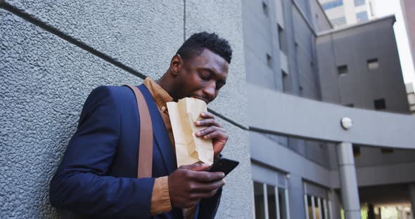 African american businessman eating sandwich using smartphone in city street alt