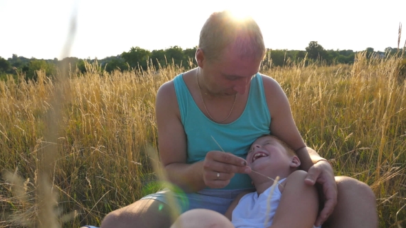 Happy Young Man Sitting on Grass at the Field and Playing with His Baby Boy alt