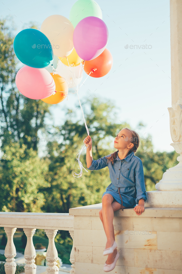 Girl holding colorful balloons looking to them while sitting in