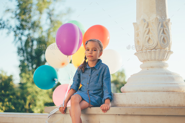 Sad girl holding colorful balloons looking to camera while sitti