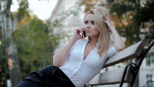 Young Beautiful Blonde Businesswoman Talking By Mobile Phone While Sitting on Benchin the City alt