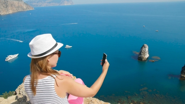 A Young Girl Does Selfie Next To the Blue Sea Sitting on a Mountain