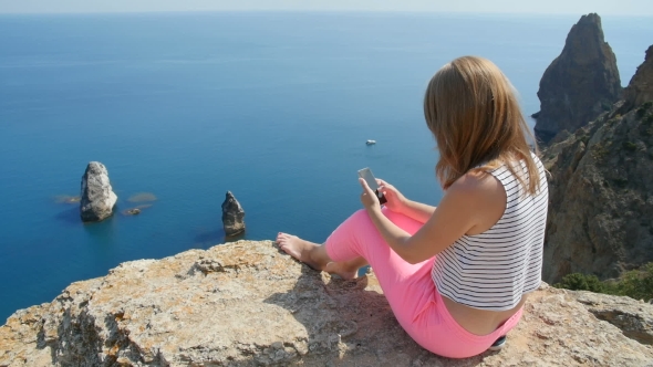 Young Girl Does Selfie Next to the Blue Sea Sitting on a Mountain
