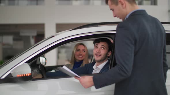 Buying Car Happy Man with Family Sitting Behind Wheel of a New Auto and Talking to Dealer in