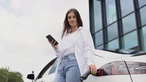 Young Woman is Standing Near the Electric Car and Looks at the Smart Phone alt