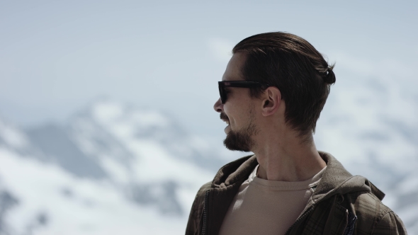 Young Bearded Man in Sunglasses at Mountain Top with Scenic View Looks Around