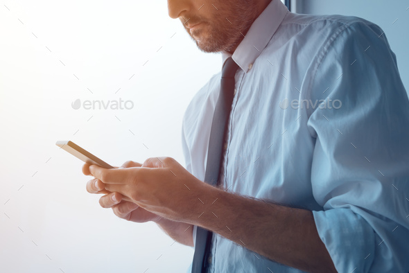 Businessman standing next to office window and texting - Stock Photo - Images