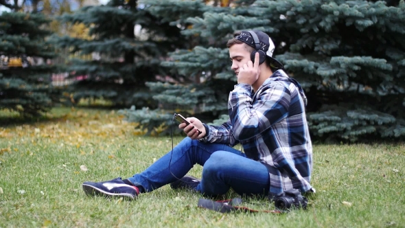 Young Male in Headphones Sitting on Grass and Listening Music