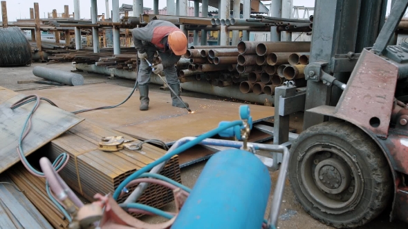 A Man in Protective Uniform and a Helmet on His Head Sawing a Large Steel Part in Order To Put It on alt