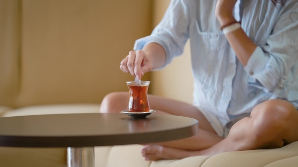 A Woman in a Man's Shirt Stirs Sugar in a Cup, a Lady Drinks Strong Tea and Sits on a Sofa alt