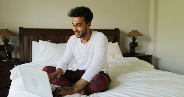 Pondering Man Using Laptop Computer Sitting On Bed Hispanic Guy Type Chatti...