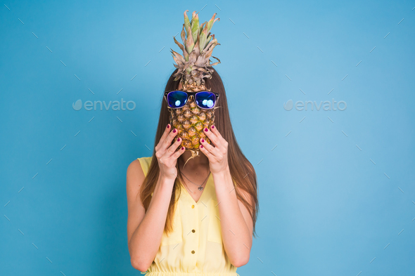 Portrait of a pretty funny summer girl holding pineapple in sunglasses isolated over blue background