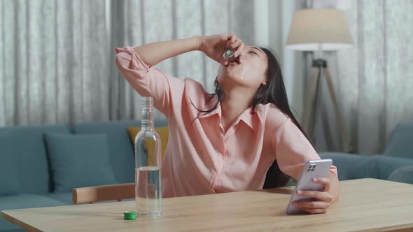 Drunk, Depressed Asian Woman Drinking Vodka From Glass During Using Smartphone At Home alt