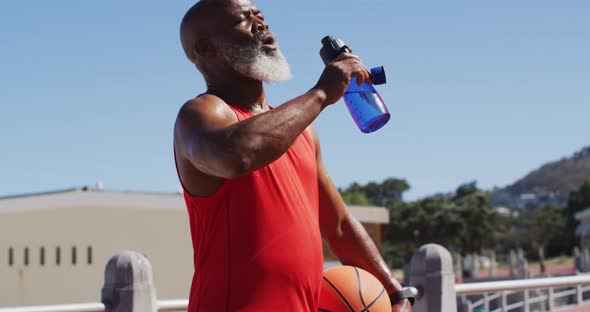 Senior african american man with basketball drinking water on the court near the beach alt