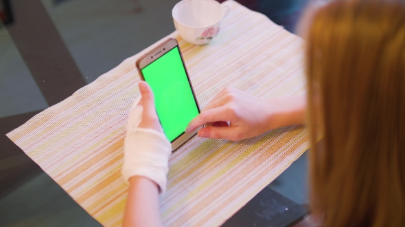 Young Caucasian Blonde Sit at Table with Broken Left Hand and Look at Green Screen of Smartphone