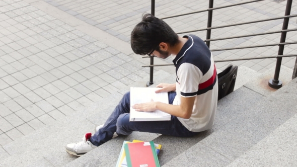 Portrait of a Happy Male Asian Student Sitting on Stairs and Reading Book