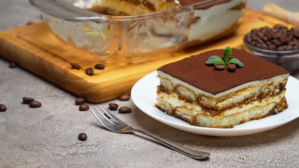 Traditional Italian Tiramisu dessert in glass baking dish and portion on grey concrete background alt