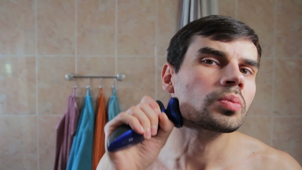 Young Man Shaving with an Electric Razor in the Bathroom. Guy Shaves His Beard with a Razor in the alt