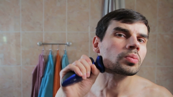 Young Man Shaving with an Electric Razor in the Bathroom. Guy Shaves His Beard with a Razor in the