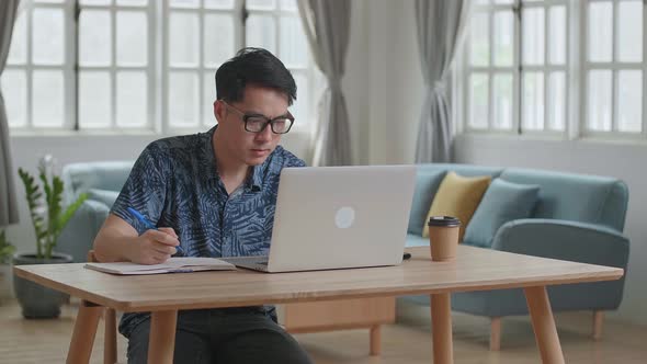Asian Man Using Laptop Computer And Writing On Notebook At Home alt