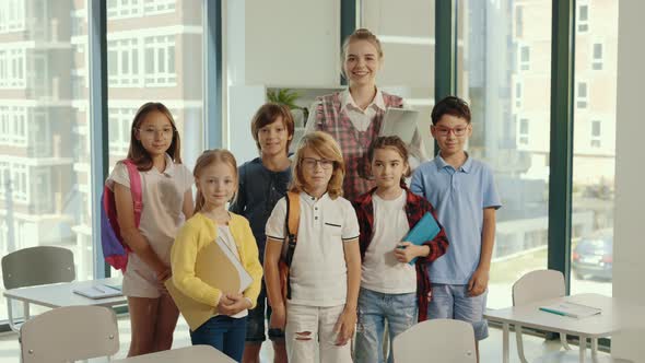 Portrait of Elementary School Pupils Standing in Classroom with Female Teacher in the Middle of the alt