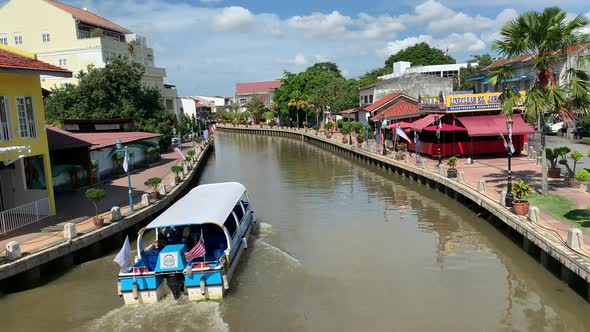 Narrow looking Malacca River with houses  while boat is cruising the river and a cloudy blue sky in alt