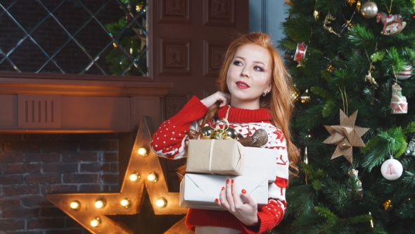 Cute Redhead Girl in the Red Sweater Near Christmas Tree with Gifts in Hand. alt